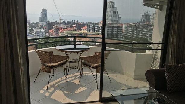 Un balcon agrémenté de fauteuils en osier et d’une table en verre, d’où l’on peut admirer un paysage urbain composé de différents bâtiments et de grues de construction.