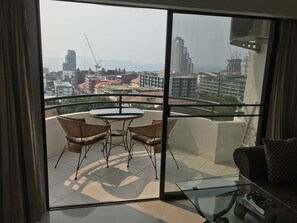 Un balcón con sillas de mimbre y una mesa de vidrio, con vistas a un paisaje urbano con varios edificios y grúas de construcción.