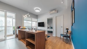 A modern kitchen with a wooden island, a dining area with a chair and table, a TV mounted on the wall, and a balcony with a view of the city.