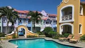 Una piscina con sillas de descanso, un edificio amarillo con balcones y edificios coloridos al fondo.