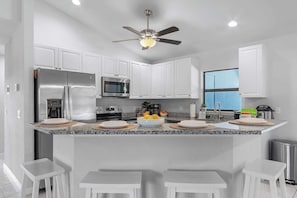 Una cocina moderna con armarios blancos, electrodomésticos de acero inoxidable, encimera de granito y ventilador de techo.
