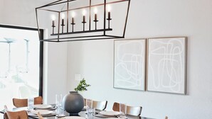 Een eetgedeelte met een rechthoekige tafel voor zes personen, omringd door houten stoelen. Daarboven een geometrische hanglamp met meerdere kaarsen. Op de muur hangen twee abstracte schilderijen.