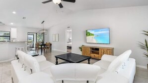 Een ruime woonkamer met een grote flatscreen-tv, witte leren banken, een houten entertainmentkast en een plafondventilator.