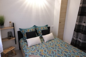 Une petite chambre à coucher, bien rangée, avec un lit, des oreillers, une étagère avec une plante et un petit tabouret.