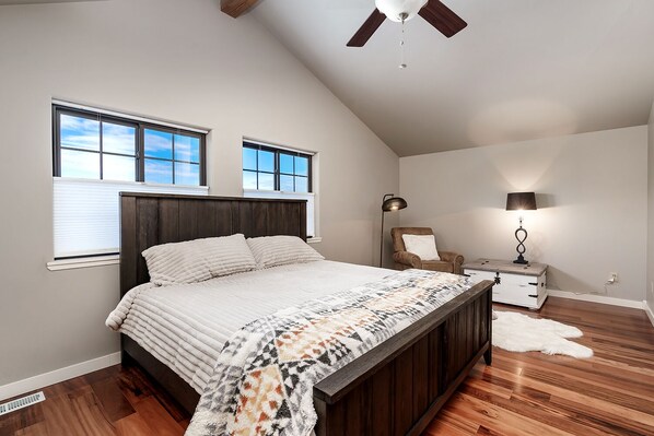 A bedroom with a large bed, two windows, a ceiling fan, a chair, a nightstand, and a lamp. - Cowboy Cabin on Cooper (Red Lodge)