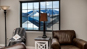 A living room with a view of mountains through a large window, a brown leather chair, a white side table, and a floor lamp. - Cowboy Cabin on Cooper (Red Lodge)