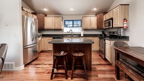 A modern kitchen with wooden cabinets, stainless steel appliances, a central island with stools, and a window providing natural light. - Cowboy Cabin on Cooper (Red Lodge)