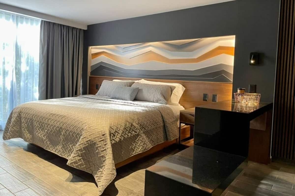 A modern hotel room with a large bed, a nightstand, a desk with a lamp, and a view of the city through the window.