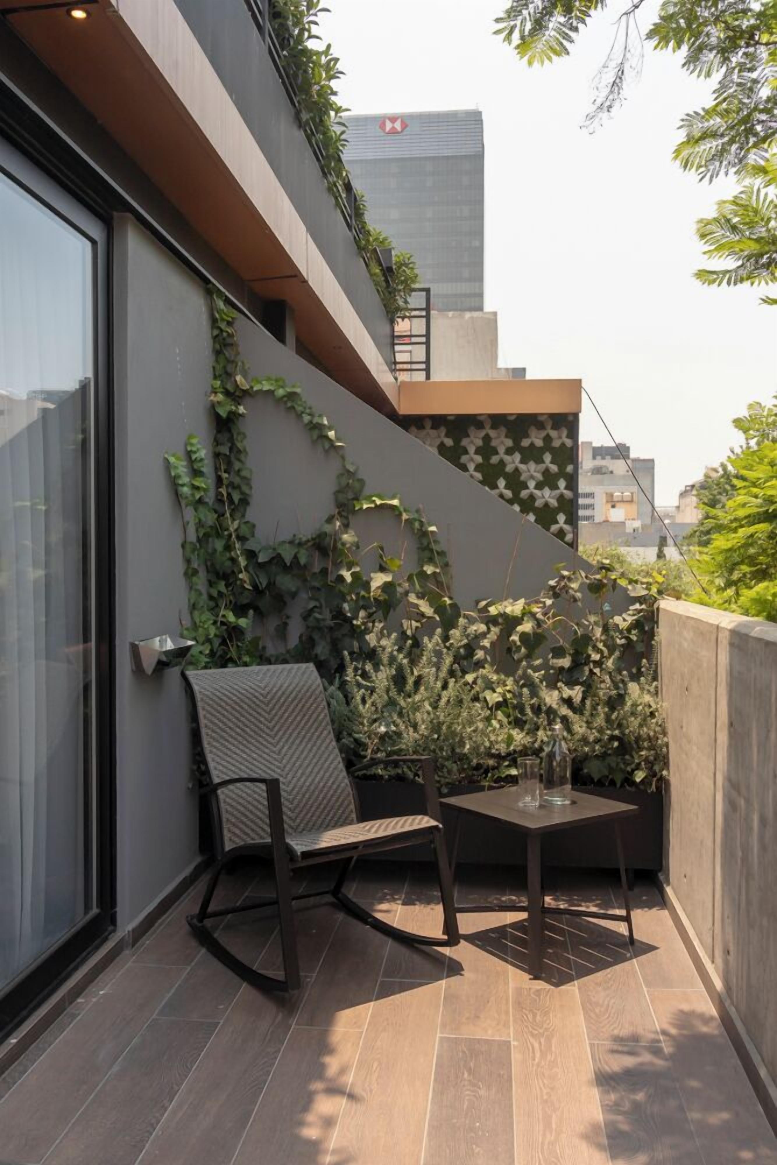 A modern balcony with a chair and table, surrounded by greenery and a patterned wall.