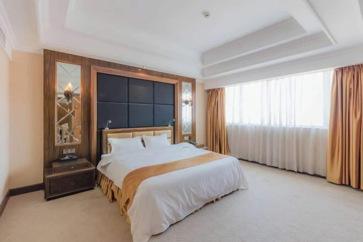A hotel room with a large bed, a wooden headboard, a nightstand with a lamp, a mirror, and a window with curtains.