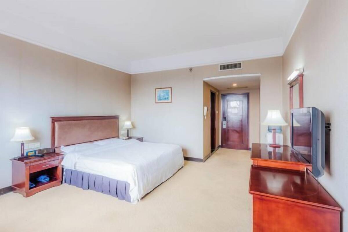 A hotel room with a large bed, a desk with a lamp, a television, and a door leading to another room.
