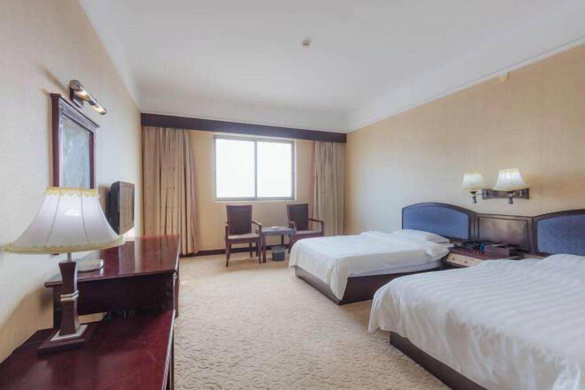 A hotel room with two beds, a desk with a lamp, a chair, a window with curtains, and a nightstand with lamps.