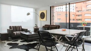 Un salon moderne comprenant un canapé, une table basse et des fauteuils, une table à manger avec des chaises, une grande fenêtre donnant sur des immeubles et un tapis en peau de vache.