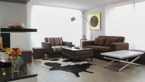 Un salon moderne comprenant un canapé, un pouf, une table basse et une table blanche. Un tapis en peau de vache recouvre le sol.