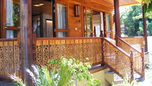 Eine hölzerne Veranda mit aufwendiger Geländerkonstruktion, einer Treppe und Blick ins Innere durch offene Türen.