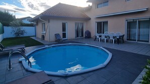 Ein Schwimmbecken mit Fliesen umgeben, eine überdachte Terrasse mit Sitzgelegenheiten und ein Gebäude mit einem Ziegeldach.