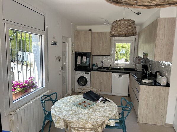 A compact kitchen with a washing machine, dishwasher, and a dining area with a table and chairs. - Charming, renovated house—4-minute walk to Grifeu Beach—Beautiful garden—Parking—Air conditioning (Llançà)