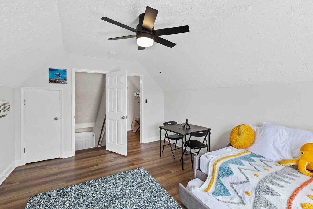 A bedroom with a bed, a small table, a ceiling fan, and a door leading to another room.