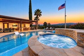 Ein Swimmingpool mit Liegestühlen, einem Whirlpool und einer Terrasse mit Blick auf den Sonnenuntergang und die Berge.