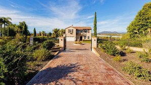 Una casa de dos pisos con un camino de entrada pavimentado que conduce a un acceso con portón, rodeada de exuberante vegetación y un cielo azul despejado.