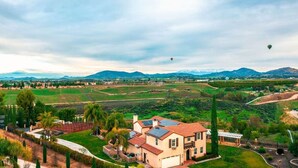 Una casa grande con techo de tejas rojas, rodeada de campos verdes y montañas al fondo.