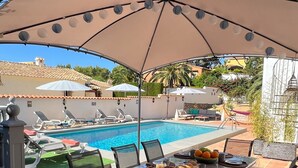 Un área de piscina al aire libre con una mesa de comedor cubierta, sillas de descanso y vistas a la piscina.