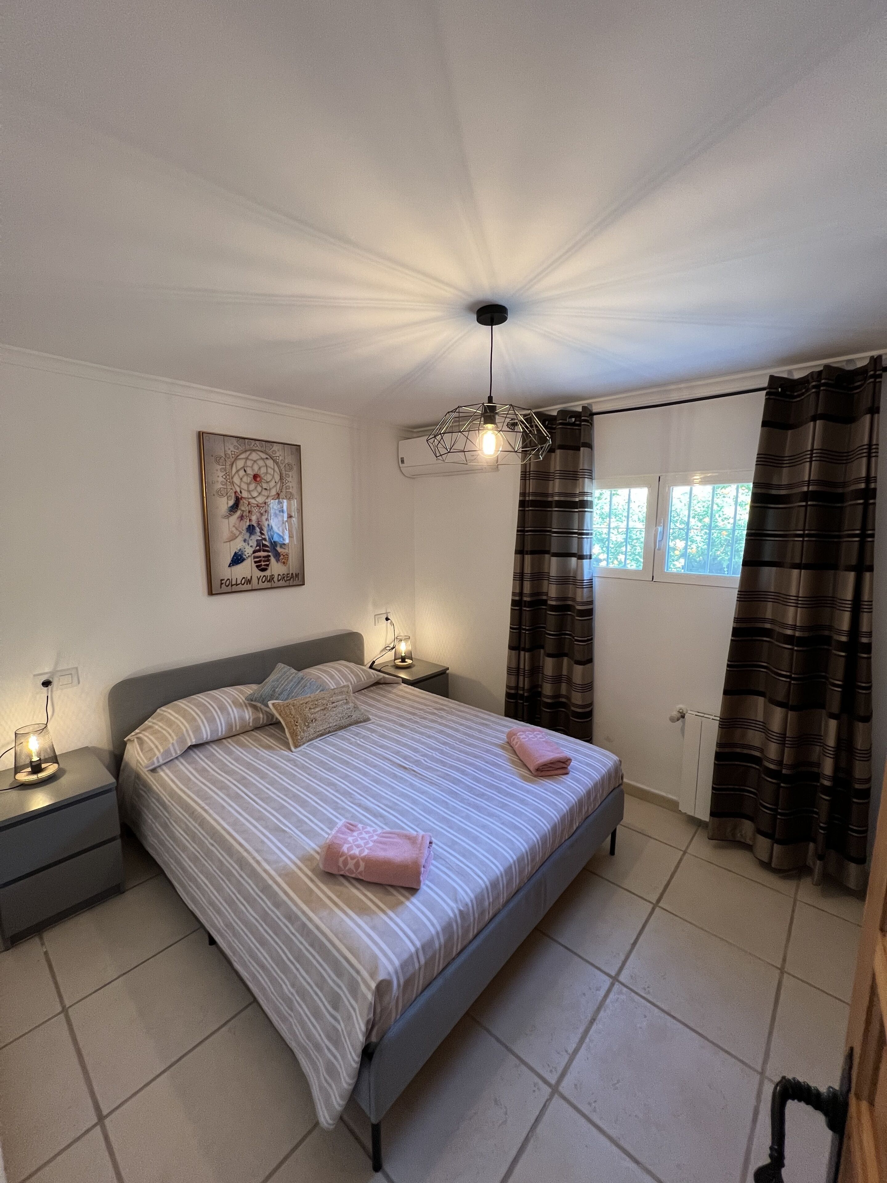 A bedroom with a bed, nightstands, a wall-mounted lamp, a window with curtains, and a ceiling light fixture.