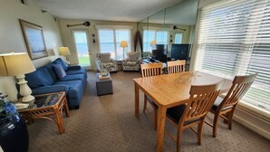 A living room with a blue sofa, a wooden dining table, and chairs. There is a TV on a stand, a lamp, and a large mirror on the wall. - Cozy Beachfront, Amazing Views - North Shore Inn Unit 106 (Traverse City)