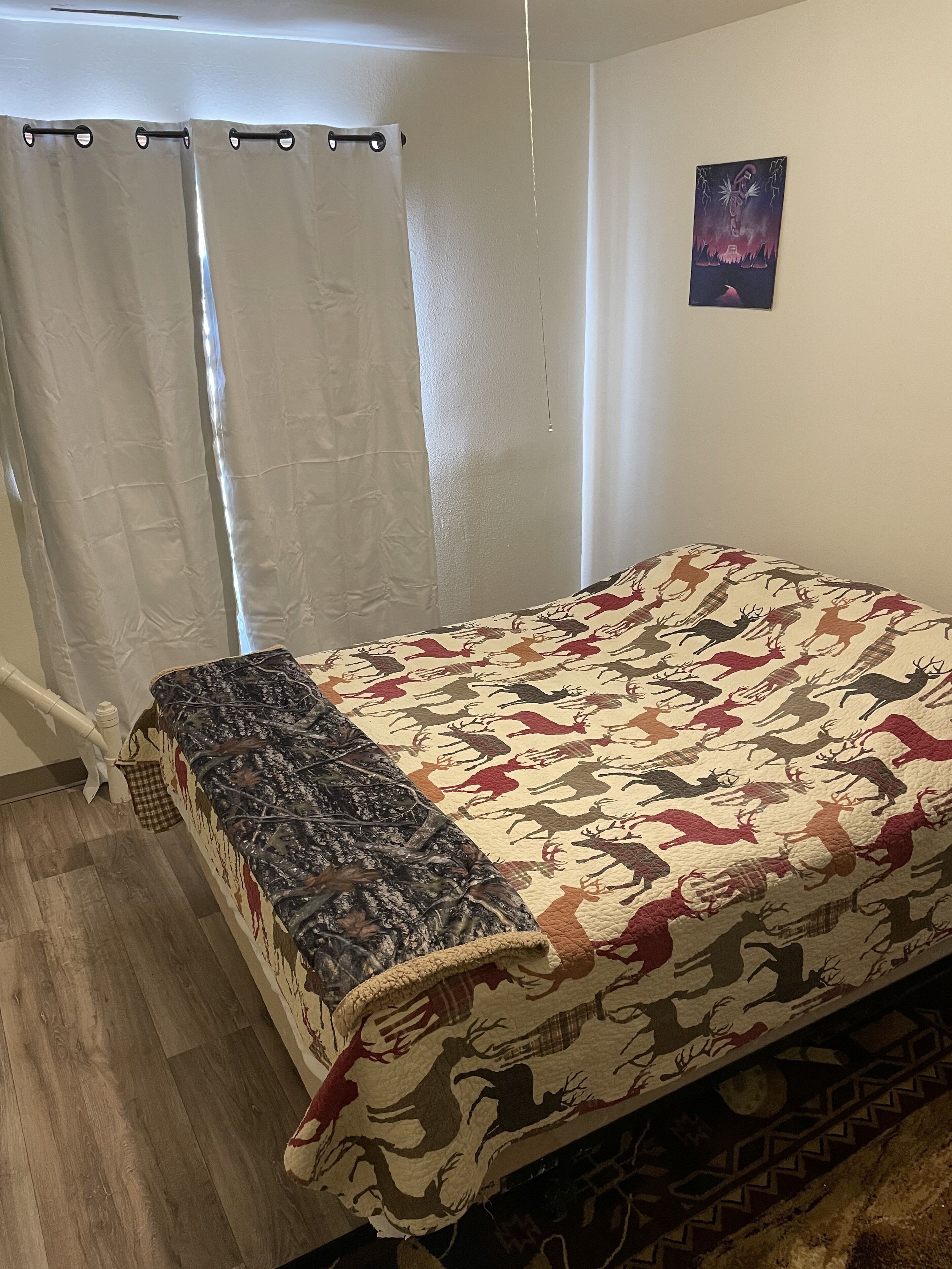 A bedroom with a bed, a window with curtains, a wall with a painting, and a wooden floor.