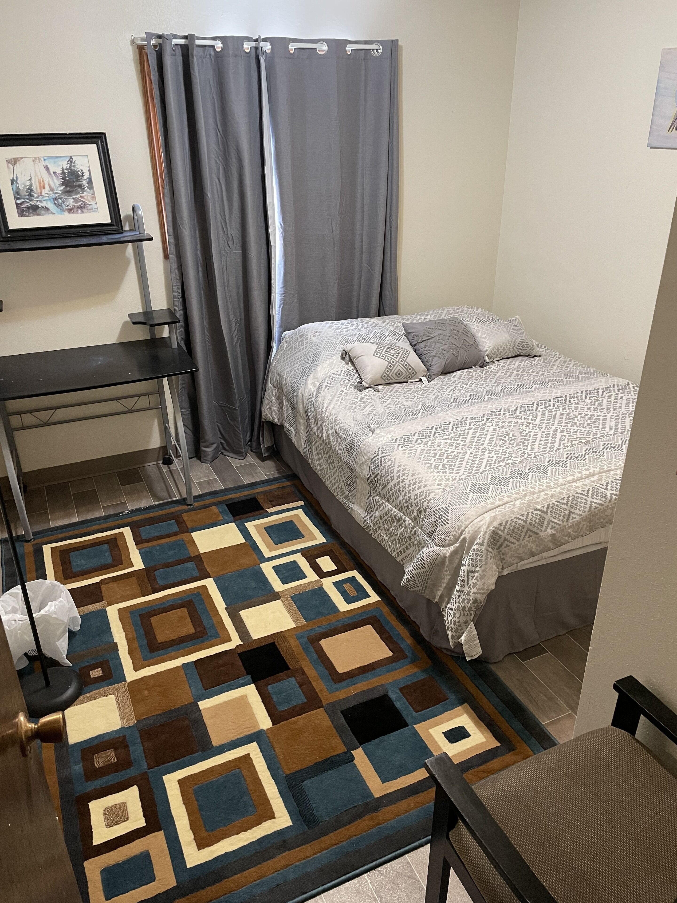 Un dormitorio con una cama, un escritorio, una silla, una alfombra y un cuadro en la pared.