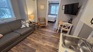 Un espacio habitable compacto con sofá, una pequeña mesa con lámpara, una silla de mimbre, zona de comedor con sillas, cama, televisor montado en la pared y cocina con fregadero.