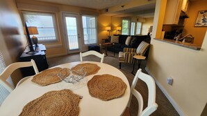 Un comedor con una mesa redonda, cuatro individuales tejidos, sillas blancas y una mesita auxiliar negra con una lámpara.