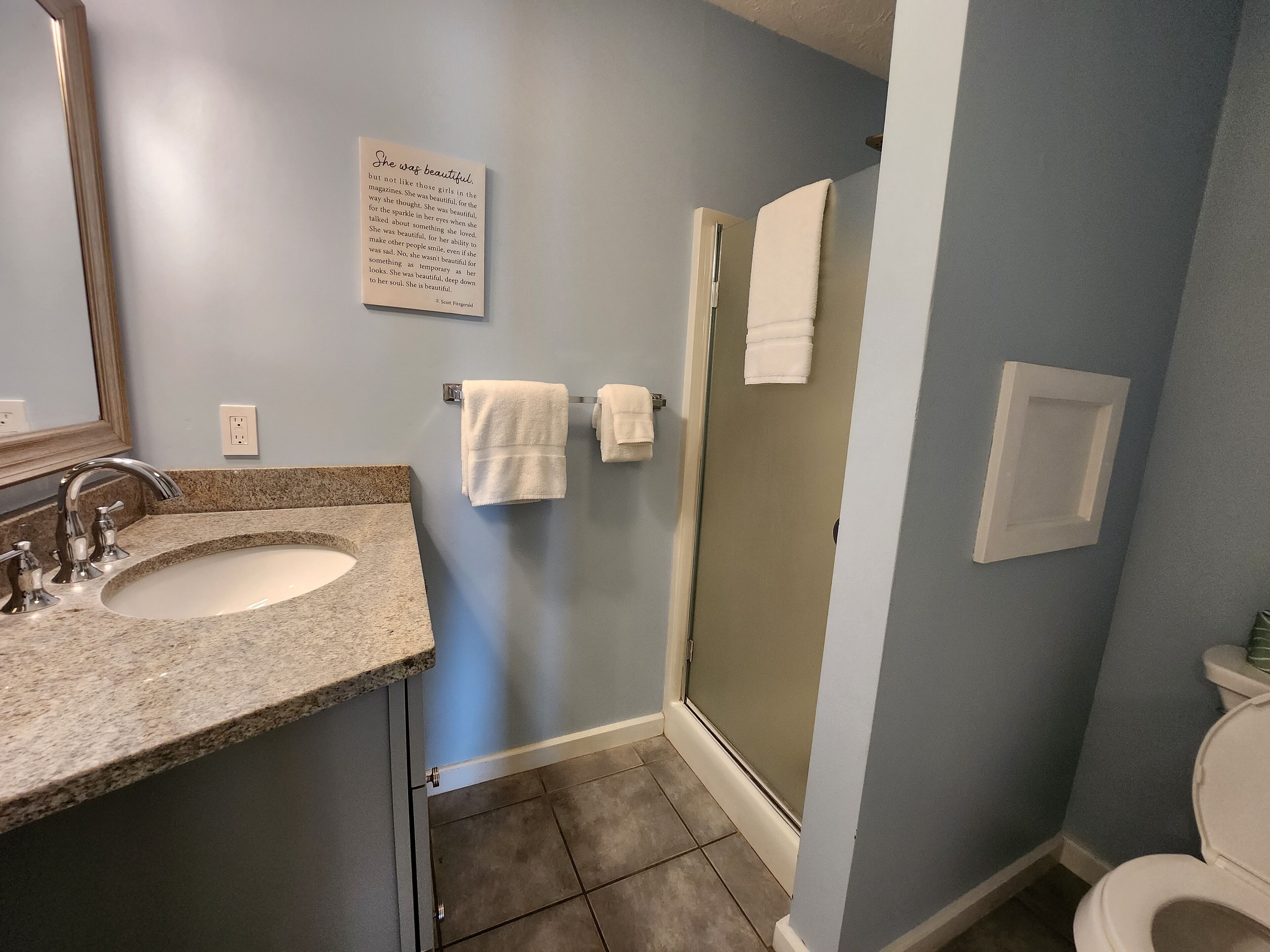 Une salle de bain avec un lavabo surmonté d’un plan de travail en granit, un miroir, une douche avec une porte en verre et des toilettes.