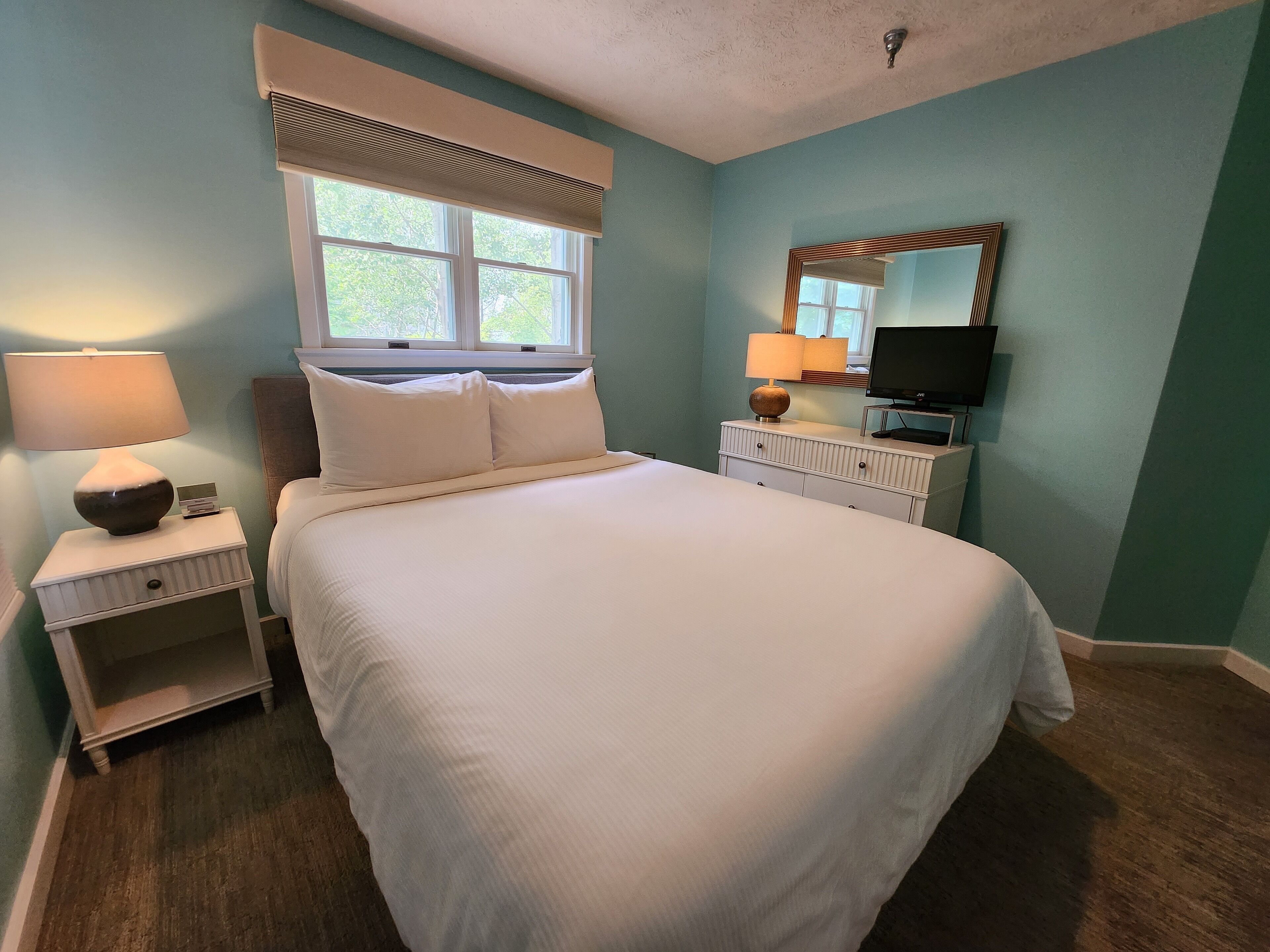 A bedroom with a bed, nightstands, a lamp, a television, and a window with blinds.