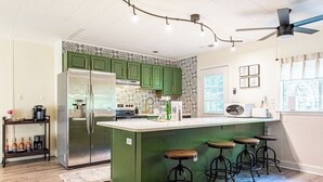 Eine Küche mit grünen Schränken, einem Edelstahlkühlschrank, einer zentralen Kochinsel mit Sitzgelegenheiten und einem Deckenventilator.