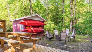 Eine Holzterrasse mit Picknicktisch und -stühlen, eine kleine Gartenhütte mit roten Kissen und ein bewaldeter Hintergrund.
