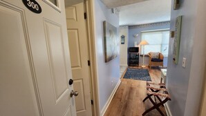 A hallway with a wooden floor, a white door with a number plate, a lamp, a wicker chair, and a small table. - Charming 1 bedroom - North Shore Inn Condo 300 (Traverse City)