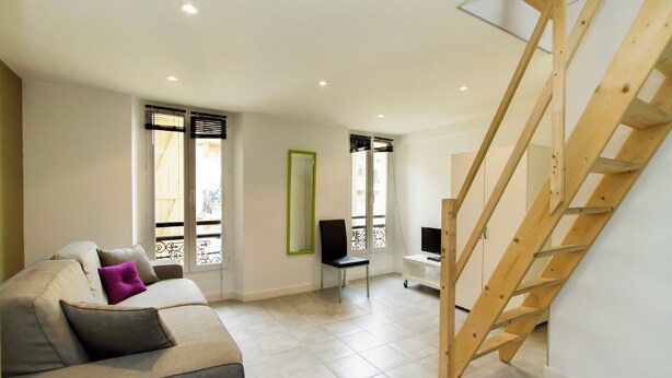 A modern living room with a sofa, a chair, a TV, and a wooden staircase. - Florella Residences 2 pièces duplex Achard (Cannes)