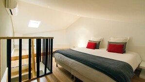 A bedroom with a bed, a nightstand, a red pillow, a white pillow, and a wooden staircase. - Florella Residences 2 pièces duplex Achard (Cannes)