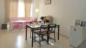 Un comedor con una mesa y sillas, un refrigerador pequeño, un sofá con funda floral rosa y una ventana con cortinas.