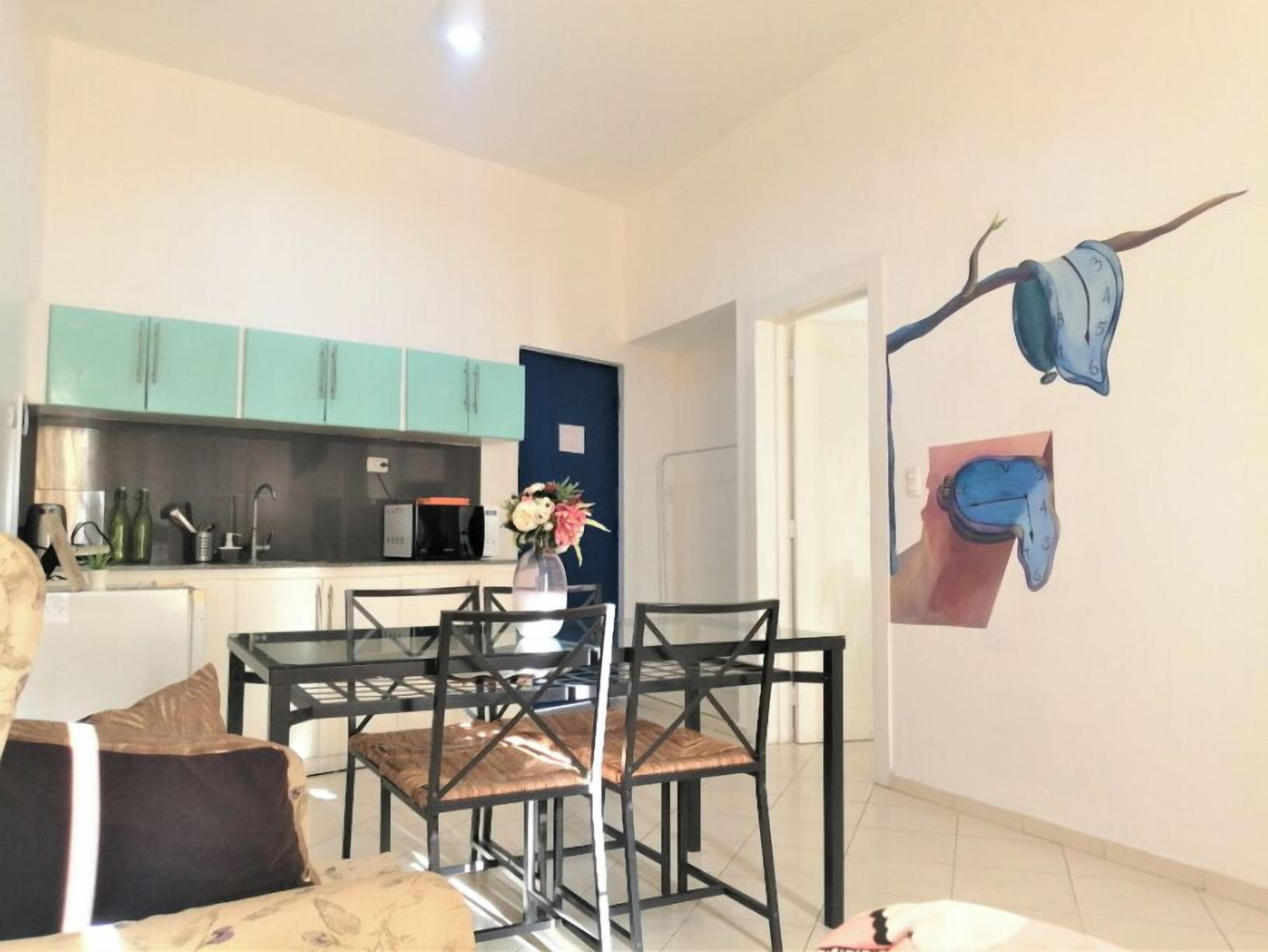 Una sala moderna con una mesa de comedor de vidrio y sillas, un sofá, y una cocina con armarios verde azulado y un microondas.