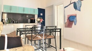 Una sala moderna con una mesa de comedor de vidrio y sillas, un sofá, y una cocina con armarios verde azulado y un microondas.