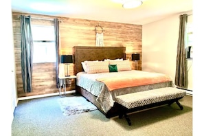 A bedroom with a wooden headboard, a bed with a patterned bedspread, two bedside tables with lamps, and a bench at the foot of the bed.