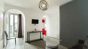 A modern living room with a grey sofa, a round coffee table, a TV mounted on the wall, and a view of the city through an arched window. - Florella Residences 2 pièces supérieur République (Cannes)