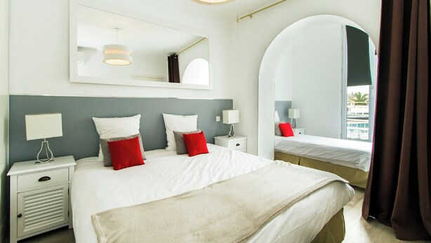 A hotel room with a large bed, a bedside table with a lamp, a mirror, and a view of the outside. - Florella Residences 2 pièces supérieur République (Cannes)