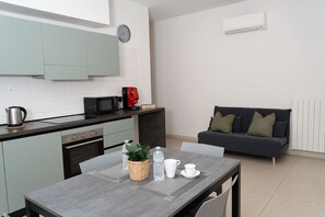 A compact kitchen with a microwave, kettle, and coffee machine, a dining table with a plant and cups, and a grey sofa in a living area.