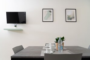 A modern dining area with a flat-screen TV mounted on the wall, a small potted plant, and a table set with white cups, saucers, and a water bottle.