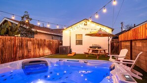 A backyard with a hot tub, string lights, a wooden fence, and a patio area with chairs and a table.