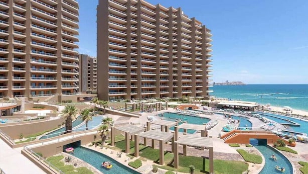 Un hotel di grandi dimensioni con numerosi balconi, una piscina centrale con scivolo acquatico e vista sulla spiaggia.