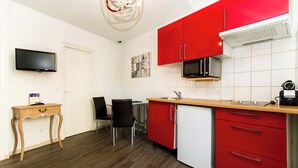 Una cocina compacta con armarios rojos, un microondas y un refrigerador pequeño. Hay una mesa de madera con sillas, un televisor de pantalla plana y un mueble bajo con cajones.
